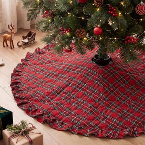 May include: A round, quilted Christmas tree skirt featuring a red, green, and white plaid pattern. The fabric skirt has a ruffled edge and is positioned at the base of a decorated evergreen tree with red ornaments and pinecones.