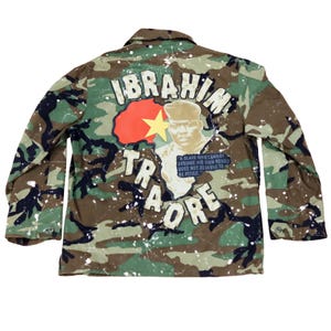 Könnte beinhalten: Tarnjacke mit dem Namen "IBRAHIM TRAORE" in weißen Buchstaben. Ein Porträt eines Mannes ist abgebildet, zusammen mit einer rot-gelben Grafik. Die Jacke hat weiße Farbspritzer.