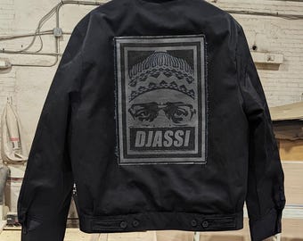 Puede incluir: Chaqueta de trabajo negra con un gran parche blanco y negro en la espalda. El parche presenta a una persona con un sombrero y gafas con la palabra "DJASSI" impresa debajo.