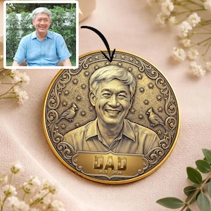 May include: A gold-colored commemorative coin featuring a smiling man's portrait, with the word "DAD" and the years "1953-2025" engraved on it. The coin has a decorative border with stars and two bird illustrations.