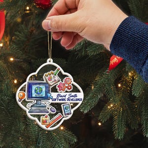 Op de afbeelding: Een witte, vierlobbige kerstversiering met software-ontwikkelaar graphics. De versiering bevat een computer, tandwielen, een notitieblok en de tekst "David Smith SOFTWARE DEVELOPER". Het wordt door een hand vastgehouden tegen een kerstboom.