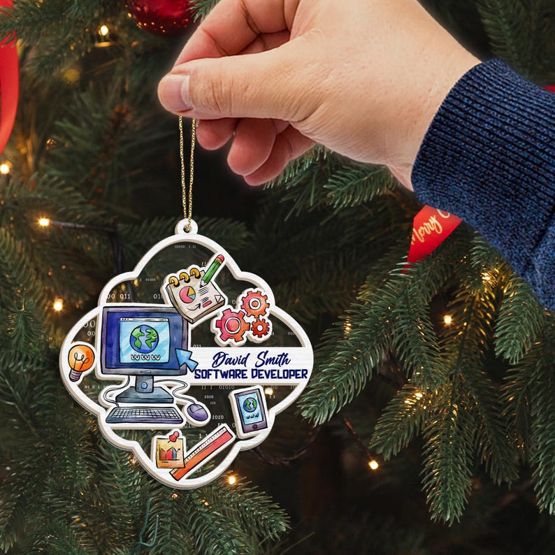 Op de afbeelding: Een witte, vierlobbige kerstversiering met software-ontwikkelaar graphics. De versiering bevat een computer, tandwielen, een notitieblok en de tekst "David Smith SOFTWARE DEVELOPER". Het wordt door een hand vastgehouden tegen een kerstboom.
