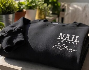 Puede incluir: Una sudadera negra con el texto "NAIL ARTIST Olivia" en blanco. La sudadera está doblada sobre una superficie de madera, con plantas verdes en el fondo.