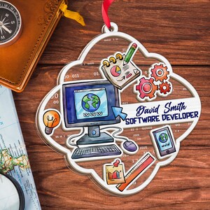 Op de afbeelding: Transparante, wolk-vormige ornament met illustraties van computer, tandwielen en tablet. Het ornament heeft de tekst "David Smith SOFTWARE DEVELOPER". Een rood lint is bevestigd om op te hangen.