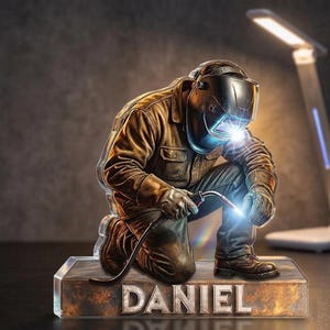 May include: A kneeling welder figurine with a welding torch, sparks, and the name "DANIEL" on the base. The welder wears a brown jacket, gloves, and a welding helmet. The base has a metallic, weathered appearance.