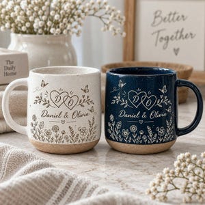 May include: Two ceramic mugs, one cream and one dark blue, both featuring a speckled finish and a tan base. Each mug displays a printed design of two interlocking hearts with the letters D and O, the names Daniel & Olivia, and floral motifs.
