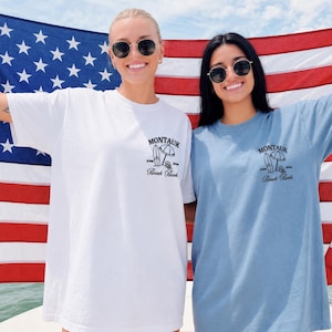 May include: A white t-shirt and a blue t-shirt, each featuring a black graphic on the left chest. The graphic depicts a surfboard, a beach umbrella, and the text 'MONTAUK', 'JUNE 2024', and 'Beach Bash'. The shirts are displayed against a large American flag.