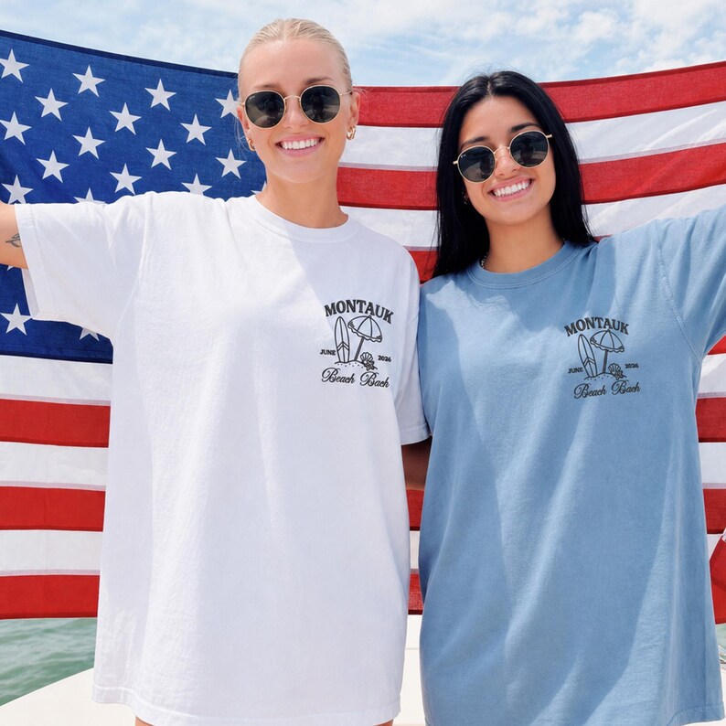May include: A white t-shirt and a blue t-shirt, each featuring a black graphic on the left chest. The graphic depicts a surfboard, a beach umbrella, and the text 'MONTAUK', 'JUNE 2024', and 'Beach Bash'. The shirts are displayed against a large American flag.