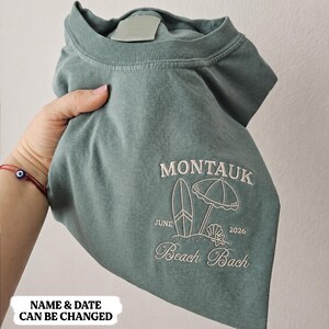 May include: A sage green crewneck t-shirt featuring white embroidery of a surfboard, a beach umbrella, a seashell, and a starfish. The text reads MONTAUK, JUNE 2026, and Beach Bach. The shirt has a ribbed collar and a blank label at the neckline.