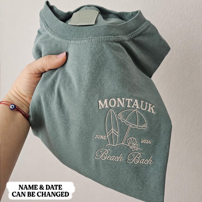 May include: A sage green crewneck t-shirt featuring white embroidery of a surfboard, a beach umbrella, a seashell, and a starfish. The text reads MONTAUK, JUNE 2026, and Beach Bach. The shirt has a ribbed collar and a blank label at the neckline.