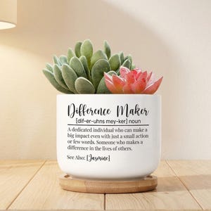 May include: A white ceramic planter with the text "Difference Maker" and a definition. The planter holds a succulent with green and red leaves, sitting on a wooden coaster. A decorative item for the home.
