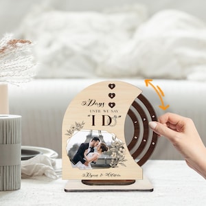May include: A wooden wedding countdown calendar featuring a photo of a couple. The calendar has a rotating dial to count down the days until the wedding. The text on the calendar reads "Days until we say I Do" and includes the couple's names.