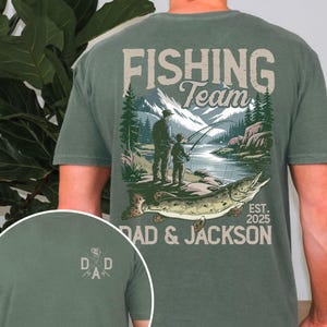 May include: Green t-shirt with a fishing scene on the back. The scene features two figures fishing in a lake with mountains in the background. The text "Fishing Team Dad & Jackson Est. 2025" is printed on the shirt.