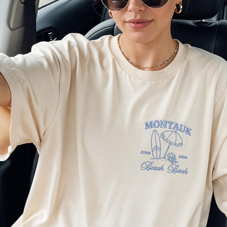 May include: A cream-colored short-sleeve t-shirt featuring light blue embroidery on the chest. The design includes the text MONTAUK, JUNE, 2026, and Beach Bash, accompanied by an embroidered surfboard, a beach umbrella, and a seashell graphic.