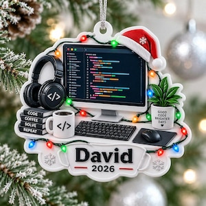 May include: A white holiday ornament featuring a computer monitor displaying code, a Santa hat, black headphones, a mug with a code symbol, a stack of books labeled Code, Coffee, Solve, Repeat, a potted plant, and the text David 2026 with string lights.