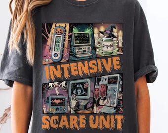 Può includere: T-shirt grigio scuro con grafica a tema Halloween. Il design presenta attrezzature mediche, un cappello da strega e le parole "Intensive Scare Unit". La maglietta ha un aspetto vintage e invecchiato.