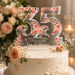 May include: A clear acrylic cake topper shaped as the number 35, decorated with a floral pattern of coral roses and green leaves. The text reads: CORAL WEDDING ANNIVERSARY, Troy & Brinna, 08-29-1991. It is placed on a white frosted cake with flowers.