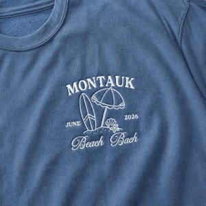 May include: A blue crewneck sweatshirt featuring white embroidered text and graphics. The design includes the word MONTAUK, a surfboard, a beach umbrella, a seashell, a starfish, the text JUNE 2026, and the words Beach Bach in script font.