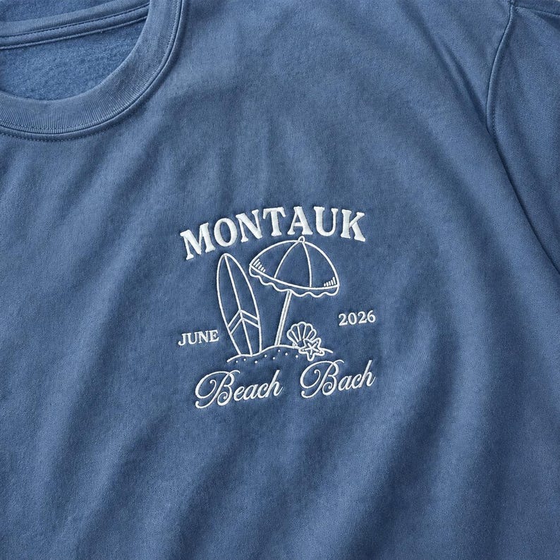 May include: A blue crewneck sweatshirt featuring white embroidered text and graphics. The design includes the word MONTAUK, a surfboard, a beach umbrella, a seashell, a starfish, the text JUNE 2026, and the words Beach Bach in script font.