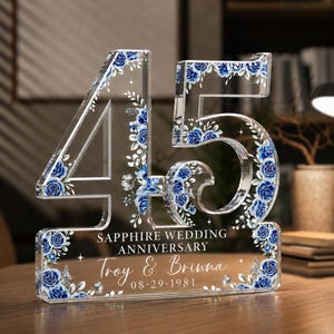 May include: A clear acrylic number 45 display featuring blue rose floral patterns. The text reads SAPPHIRE WEDDING ANNIVERSARY, Troy & Brinna, and 08-29-1981. The design includes small star accents and green leaves integrated into the floral arrangement.