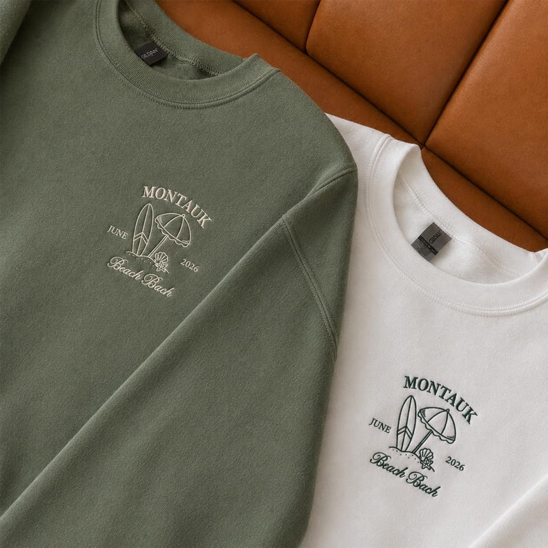 May include: An olive green sweatshirt and a white sweatshirt, each featuring an embroidered design on the chest. The design includes a surfboard, a beach umbrella, a seashell, and the text "MONTAUK JUNE 2026 Beach Bach" in a script font.