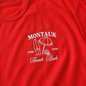 May include: A bright red sweatshirt featuring white embroidery on the chest. The design includes the text MONTAUK, JUNE 2026, and Beach Bach, accompanied by a graphic of a surfboard, a beach umbrella, a seashell, and a small starfish.