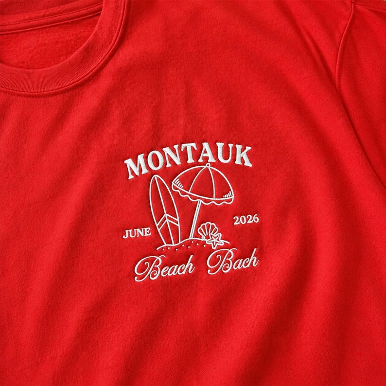 May include: A bright red sweatshirt featuring white embroidery on the chest. The design includes the text MONTAUK, JUNE 2026, and Beach Bach, accompanied by a graphic of a surfboard, a beach umbrella, a seashell, and a small starfish.