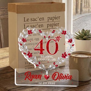 May include: A clear acrylic wedding anniversary decoration with a heart-shaped floral design and the number "40". The names "Ryan" and "Olivia" are displayed in red, along with the date 12.20.1986. A coffee mug and a succulent plant are in the background.