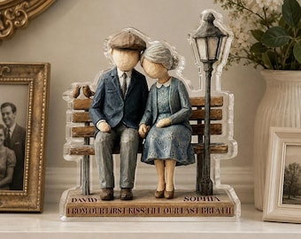 May include: A decorative figurine featuring an elderly couple sitting on a bench next to a street lamp. The man wears a blue suit and cap, the woman a blue dress. The base is engraved with the names David and Sophia and the text FROM OUR FIRST KISS TILL OUR LAST BREATH.