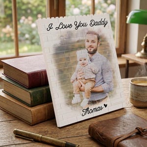 May include: A personalised photo print showing a man and a baby against a brick wall effect. The text "I Love You Daddy" and the name "Thomas" are in script. The print is on a wooden surface with books and a pen. The image is in a room.