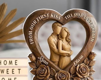 May include: A wooden heart-shaped sculpture with a couple embracing inside. The sculpture is brown with the words "FROM OUR FIRST KISS TILL OUR LAST BREATH" carved around the heart. The names "Ryan" and "Olivia" are also carved on the sculpture. Roses are carved at the base.