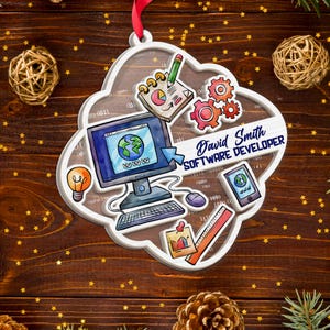 Op de afbeelding: Een witte, wolk-vormige ornament met een computer, tandwielen en een smartphone-ontwerp. Het ornament heeft de tekst "David Smith SOFTWARE DEVELOPER". Een rood lint is bevestigd om op te hangen. De achtergrond is een houten oppervlak.