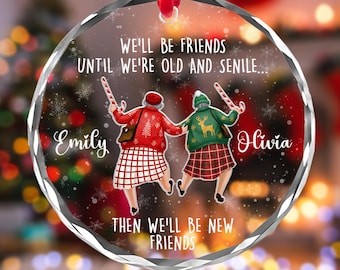 May include: A round, clear glass Christmas ornament with a red ribbon. The ornament features two elderly women holding hands, wearing sweaters and plaid skirts. Text reads "WE'LL BE FRIENDS UNTIL WE'RE OLD AND SENILE... THEN WE'LL BE NEW FRIENDS."