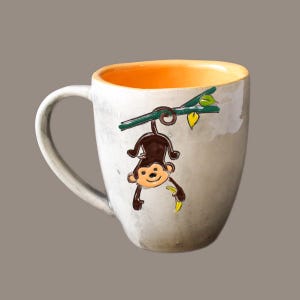May include: A white ceramic mug with an orange interior features a hand-painted design of a monkey hanging from a vine with a banana. The monkey has brown fur, a yellow banana, and green leaves.