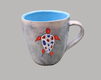 May include: A ceramic mug with a light blue interior and a grey exterior. The mug features a hand-painted turtle design in red, blue, and white. The handle is a light grey colour. Ideal for tea or coffee, this mug is handmade.