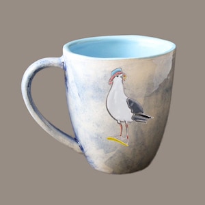 Op de afbeelding: Een keramische mok met een blauwe en witte glazuur. De mok heeft een handgeschilderde illustratie van een meeuw met een rode en blauwe hoed.