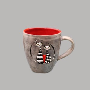 Peut inclure: Mug en céramique grise avec un intérieur rouge. La tasse présente une illustration en noir et blanc de deux chats stylisés à l'intérieur d'un cercle noir, avec un cœur rouge entre eux. La tasse a une anse incurvée.