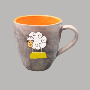 Könnte beinhalten: Graue Keramiktasse mit orangefarbenem Innenraum und weißem Schafdesign. Das Schaf hat einen lockigen Wollkörper, ein gelbes Gesicht und steht auf einem gelben Sockel. Die Tasse hat einen gebogenen Henkel und ist für Heiß- oder Kaltgetränke geeignet.