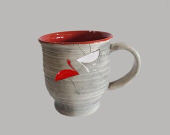 May include: A handmade ceramic mug with a gray exterior and a red interior. The mug features a large handle and is decorated with red and white abstract shapes. The mug is suitable for hot or cold beverages.