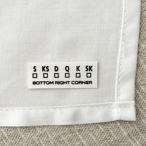 Op de afbeelding: Witte stoffen label met zwarte tekst die "S KS D Q K SK" luidt met zes lege vakjes eronder. De tekst "BOTTOM RIGHT CORNER" staat onder de vakjes.
