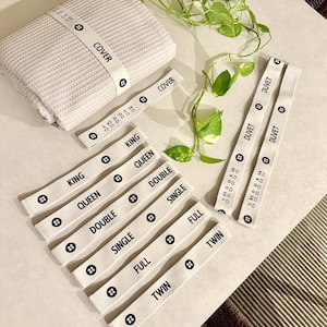 May include: A stack of light gray waffle-weave fabric with a white band labeled "COVER". Several white bands with black text indicating bed sizes like "KING", "QUEEN", "DOUBLE", "SINGLE", "FULL", and "TWIN".