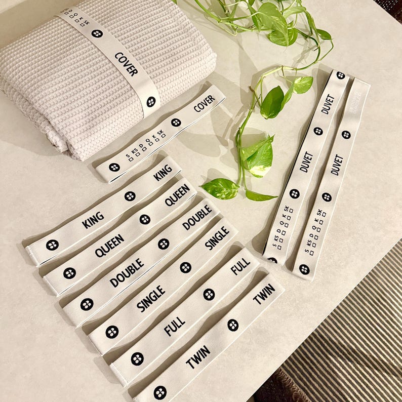 May include: A stack of light gray waffle-weave fabric with a white band labeled "COVER". Several white bands with black text indicating bed sizes like "KING", "QUEEN", "DOUBLE", "SINGLE", "FULL", and "TWIN".
