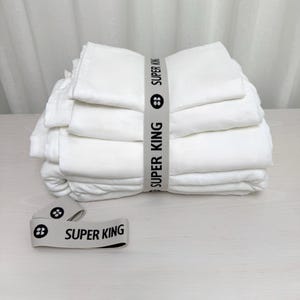 May include: A stack of folded white bed sheets, secured with a gray band. The band is printed with the words "SUPER KING" in black, along with a logo. A matching band lies in front of the stack. The sheets are neatly arranged on a light-colored surface.