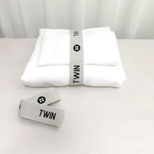 May include: A neatly folded white bedding set, secured with a gray band labeled "TWIN" in black text. Two additional tags with the same text and a symbol are placed in front. The set is on a white surface.