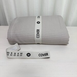 May include: A light gray waffle weave fabric with the word "COVER" printed on a white label. The fabric is folded in half and a white label with the word "COVER" and a button is attached to the fabric.