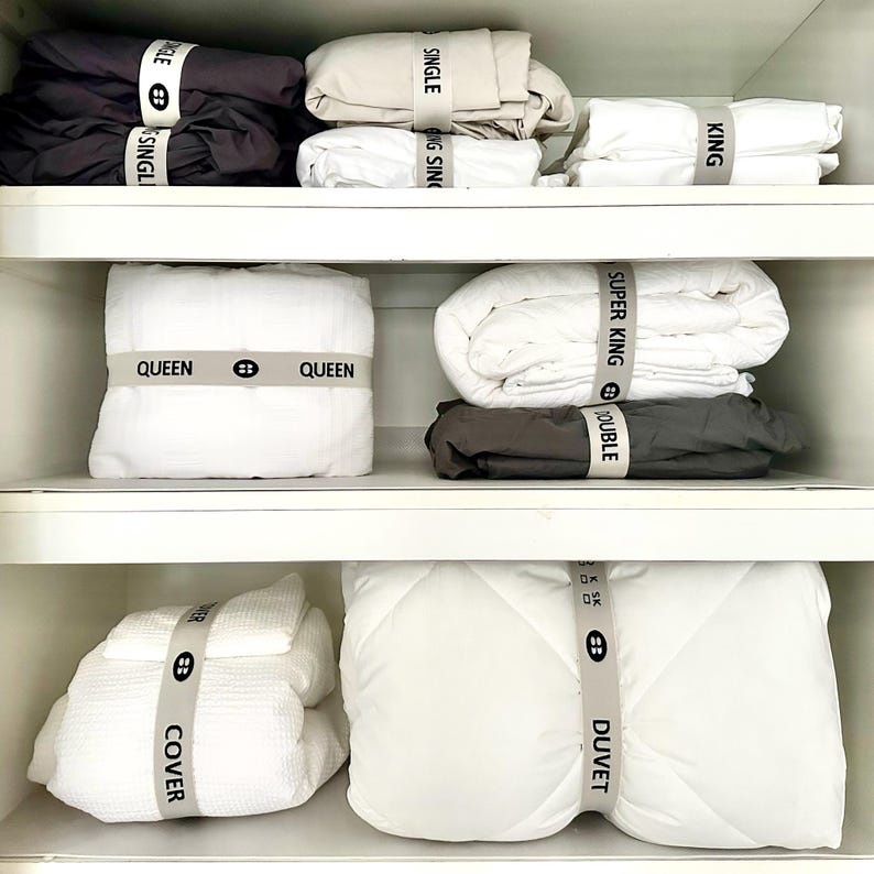 May include: A neatly organized closet displays folded bedding sets in various sizes. The sets are in neutral colors like white, beige, and charcoal, each secured with a gray band labeled with the size, such as "SINGLE" or "QUEEN". The overall aesthetic is clean and minimalist.