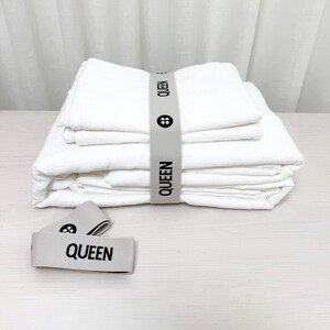 May include: A stack of white cotton bed sheets with a gray elastic band labeled "QUEEN" on each side. The sheets are folded and stacked on a white surface.