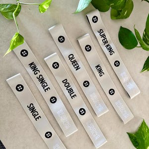 May include: Off-white fabric bed size labels with black text. Labels include "SINGLE", "KING SINGLE", "DOUBLE", "QUEEN", "KING", and "SUPER KING". Each label has a black button graphic and the "BEDDING BUNDLER" logo.