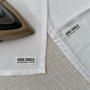 May include: White cotton sheet with a black label that reads "KING SINGLE BOTTOM RIGHT CORNER". An iron is in the background.