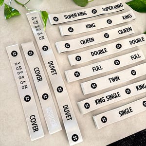 May include: White fabric bed size labels with black text. Labels include "SUPER KING", "KING", "QUEEN", "DOUBLE", "FULL", "TWIN", "KING SINGLE", and "SINGLE". Each label has a button graphic.